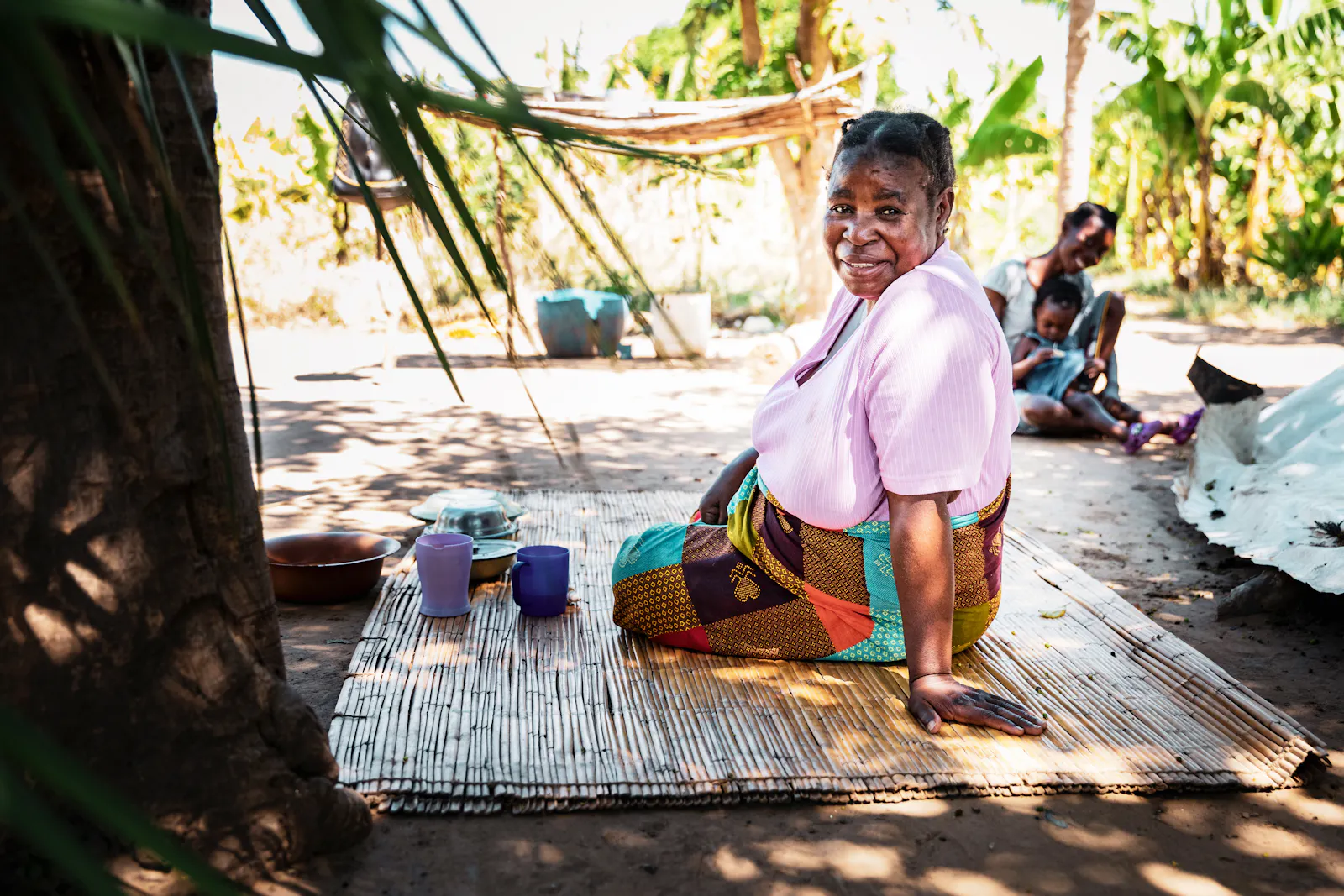 Eine bunt gekleidete afrikanische Frau sitzt auf einem Teppich und lächelt in die Kamera.