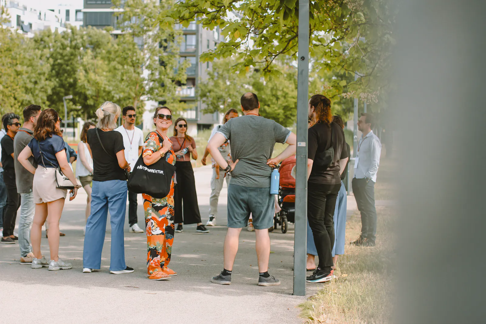Eine Gruppe von Menschen steht bei sonnigem Wetter im Schatten von Bäumen auf einem breiten Gehweg in einem modernen Stadtviertel. Im Vordergrund lächelt eine Frau in farbenfrohem, orange-schwarzem Outfit in die Kamera. Sie trägt eine schwarze Tasche mit der Aufschrift „AUTOKORREKTUR“. Die Gruppe wirkt entspannt und im Gespräch; einige Menschen halten Wasserflaschen, andere tragen Namensschilder. Im Hintergrund sind moderne Wohnhäuser und grüne Bäume zu sehen. Die Atmosphäre wirkt offen und freundlich.