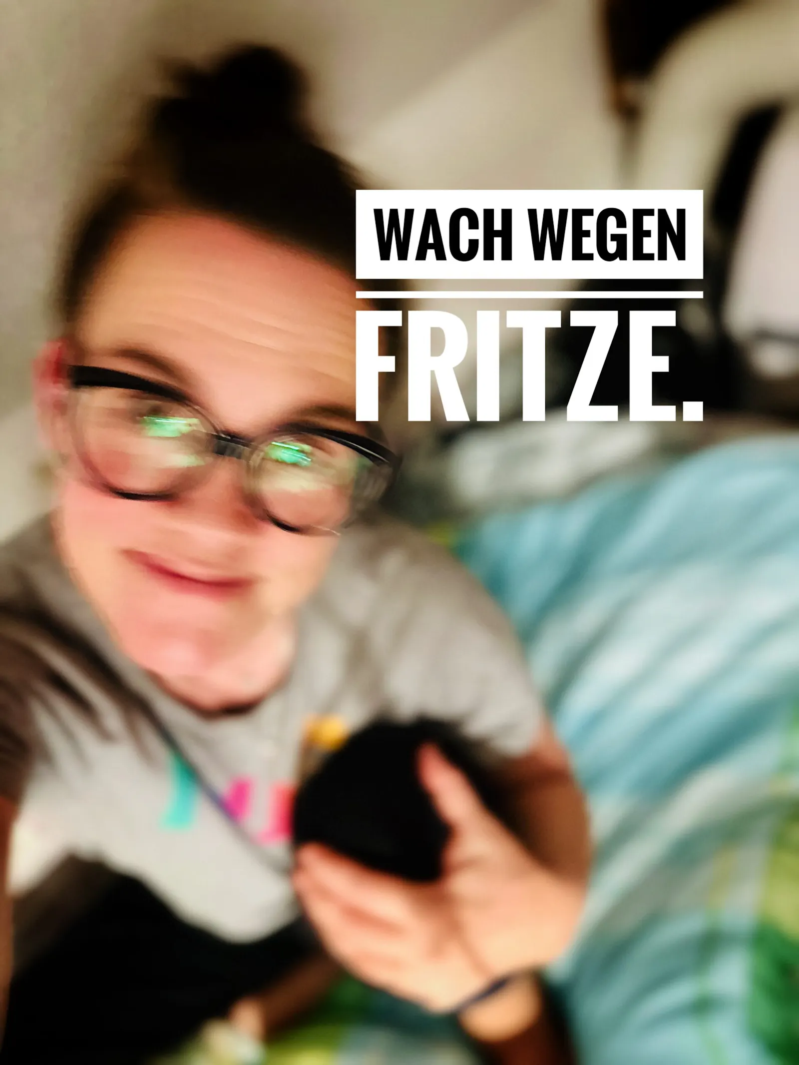 Verschwommenes Selfie einer Person mit Brille, die leicht lächelt und ein schwarzes, rundes Objekt im Arm hält. Im Bild steht in großen weißen Buchstaben: „WACH WEGEN FRITZE.“