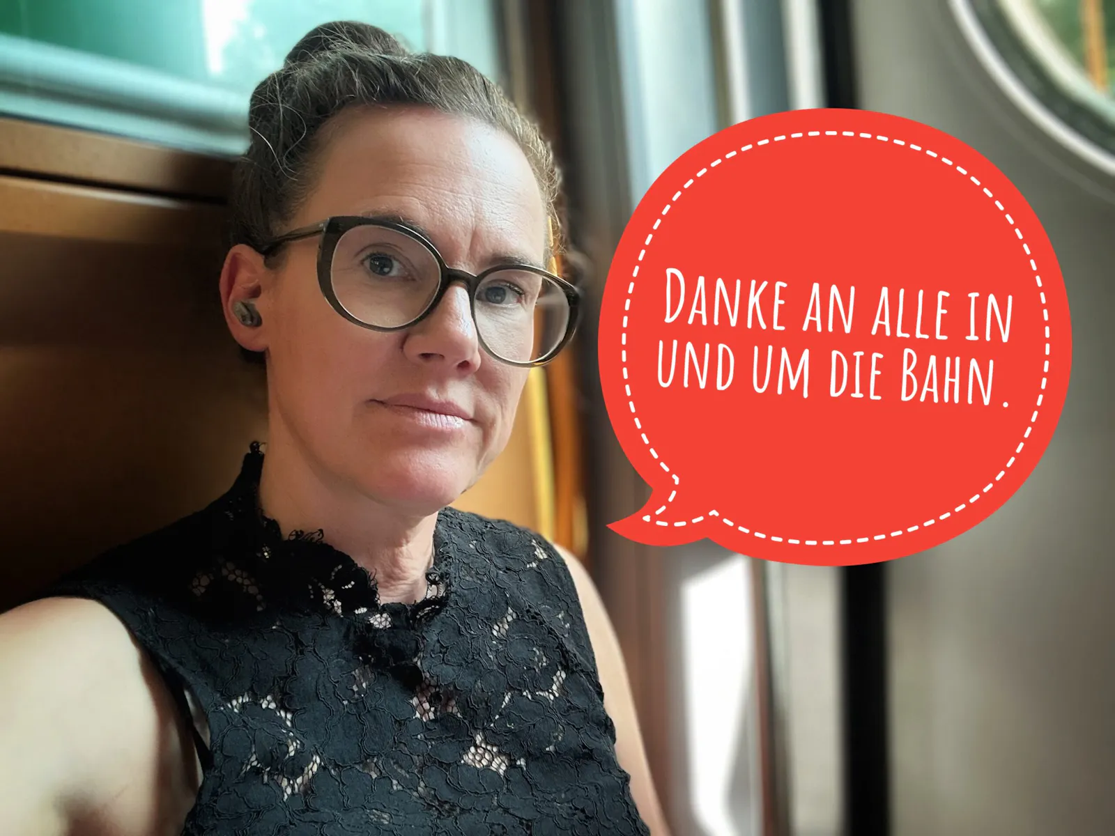 Katja sitzt an die Wand an einem Ausgang der Bahn gelehnt, in der sie fährt. Sie schaut in die Kamera und trägt ein schwarzes gesticktes Top.