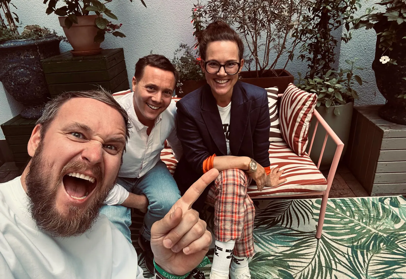 Drei Personen sitzen gemeinsam auf einer bunt gemusterten Outdoor-Couch in einem begrünten Innenhof. Links vorne im Bild hält ein Mann mit Bart die Kamera und zeigt lachend auf die beiden anderen – ein Mann in weißem Hemd und eine Frau mit Brille, Blazer und karierten Hosen, die beide freundlich in die Kamera lächeln. Im Hintergrund stehen große Pflanzen in Töpfen.