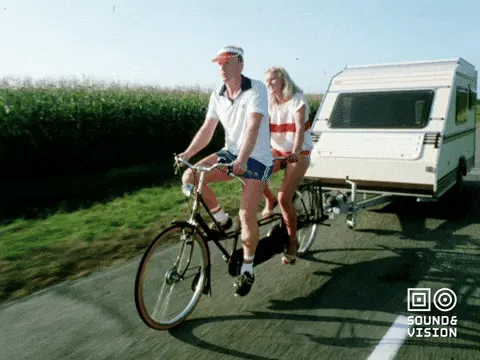 Ein Paar auf einem Tandem zieht mit dem Tandem einen kleinen Wohnwagen über eine Landstraße.