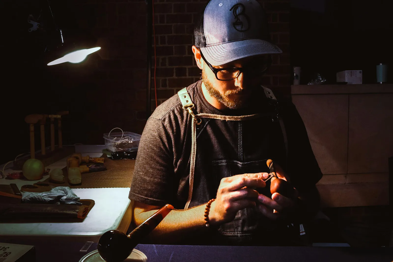 The photo of a man wearing glasses, a cap, and a work apron seated at workbench, carefully crafting a wooden pipe.