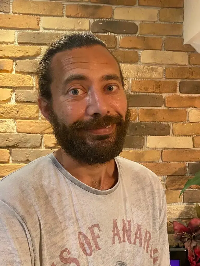 Karam Bales. Ein Mann mit kurzem braunen Haar und einem etwas buschigem braunen Bart. Er trägt ein weißes Sons of Anarchy T-Shirt und lächelt vor einer Ziegelmauer in die Kamera.