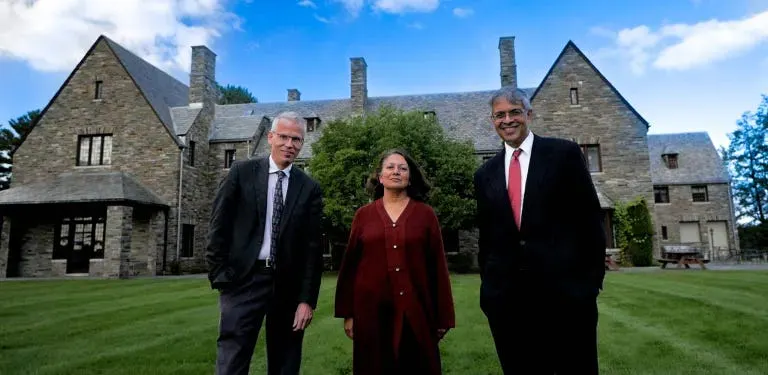 Martin Kulldorff, Sunetra Gupta, and Jay Bhattacharya stand in front of their property in Great Barrington, which boasts a green lawn. The property looks like a slightly older villa with a tree in front. They wear elegant attire and smile at the camera.