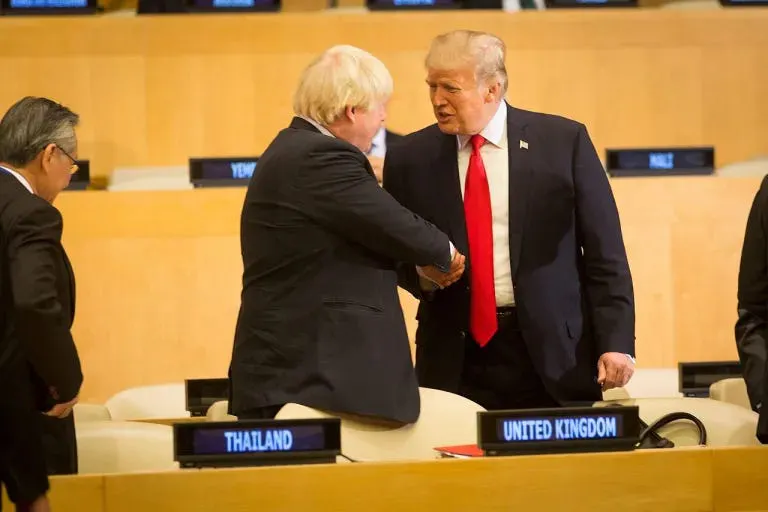 Donald Trump schüttelt die Hand von Boris Johnson in der Generalversammlung der Vereinten Nationen in New York. Sie stehen vor dem Länderschild des Vereinigten Königreich an einem langen Holztisch. Im Hintergrund weitere Sitzreihen. Links daneben das Länderschild von Thailand.