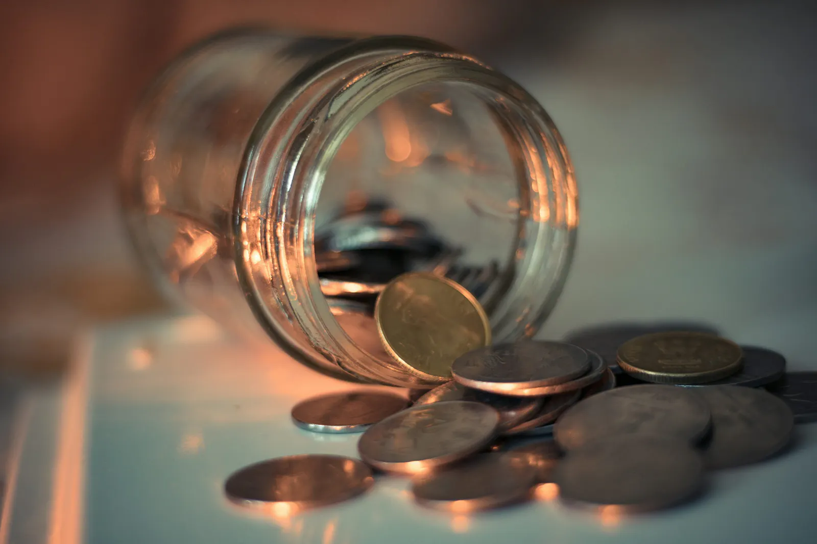 The photo of a glass jar tipped over with coins spilling out onto a white surface, lit by warm ambient light