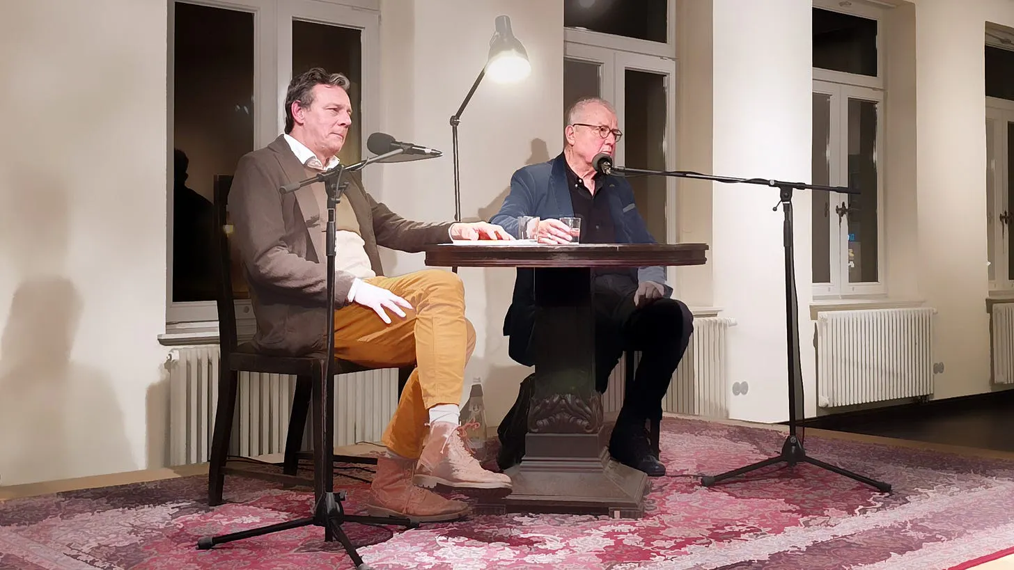 Alexander Suckel und Ruprecht Polenz sitzen nebeneinander auf einer Bühne. Vor ihnen Mikrophone, ein Tisch mit Wassergläsern. Ein roter Teppich liegt auf der Bühne. Im Hintergrund Fenster. Es ist draußen dunkel.