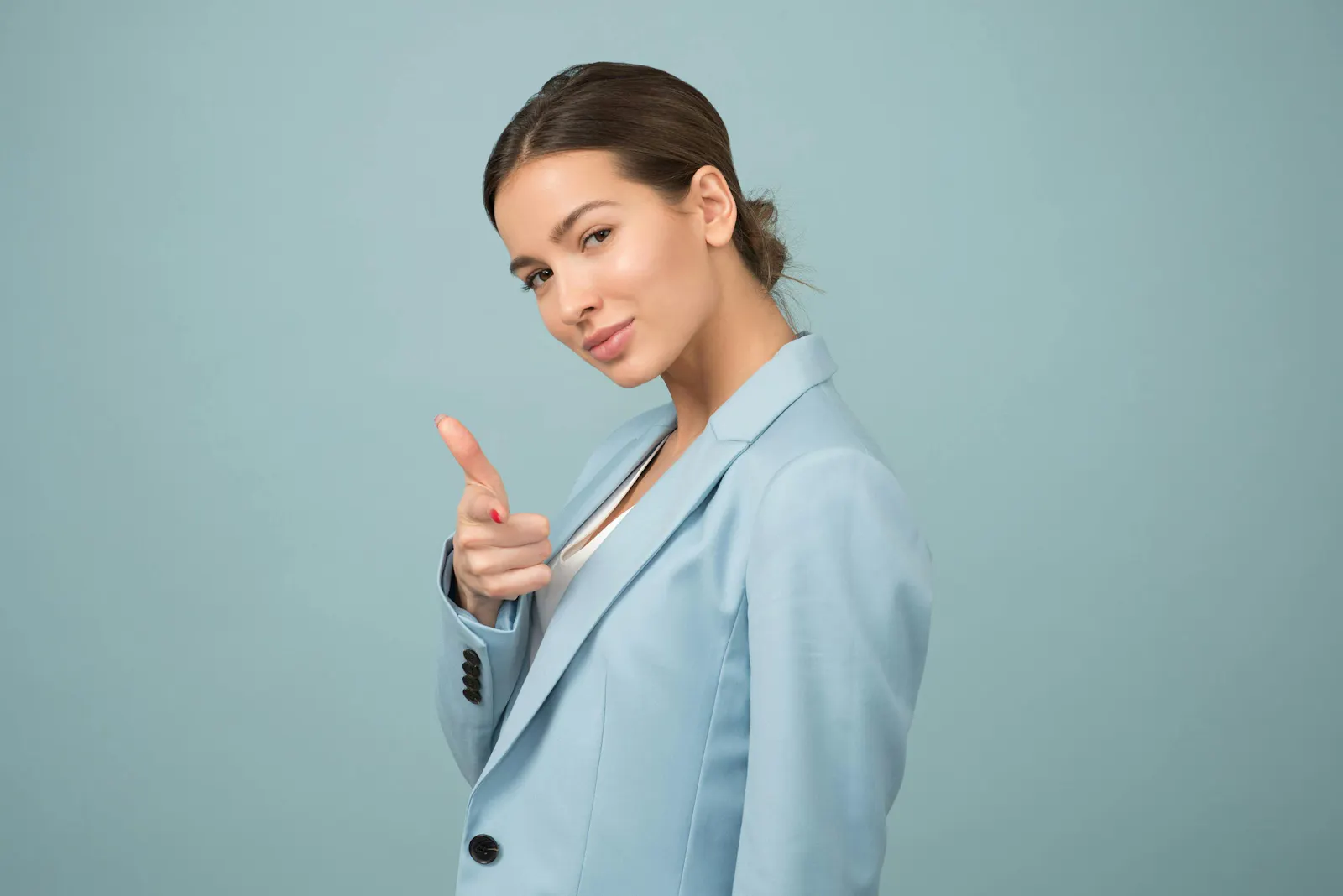 A medium close-up studio shot of a woman with her hair pulled back, wearing a light blue blazer over a white top. She is looking at the camera and pointing her right index finger forward, as if gesturing to the viewer. The background is a solid light blue.