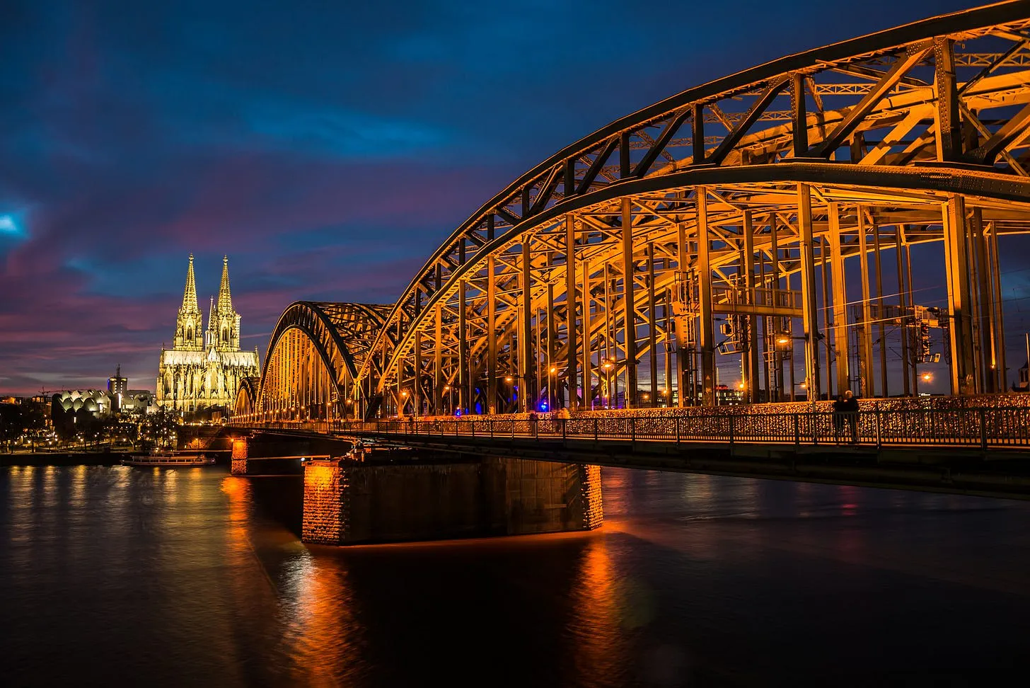 Die Brücke über den Rhein in Köln bei Abendlicht. Sie ist gelblich angestrahlt. Im Hintergrund der Kölner Dom.