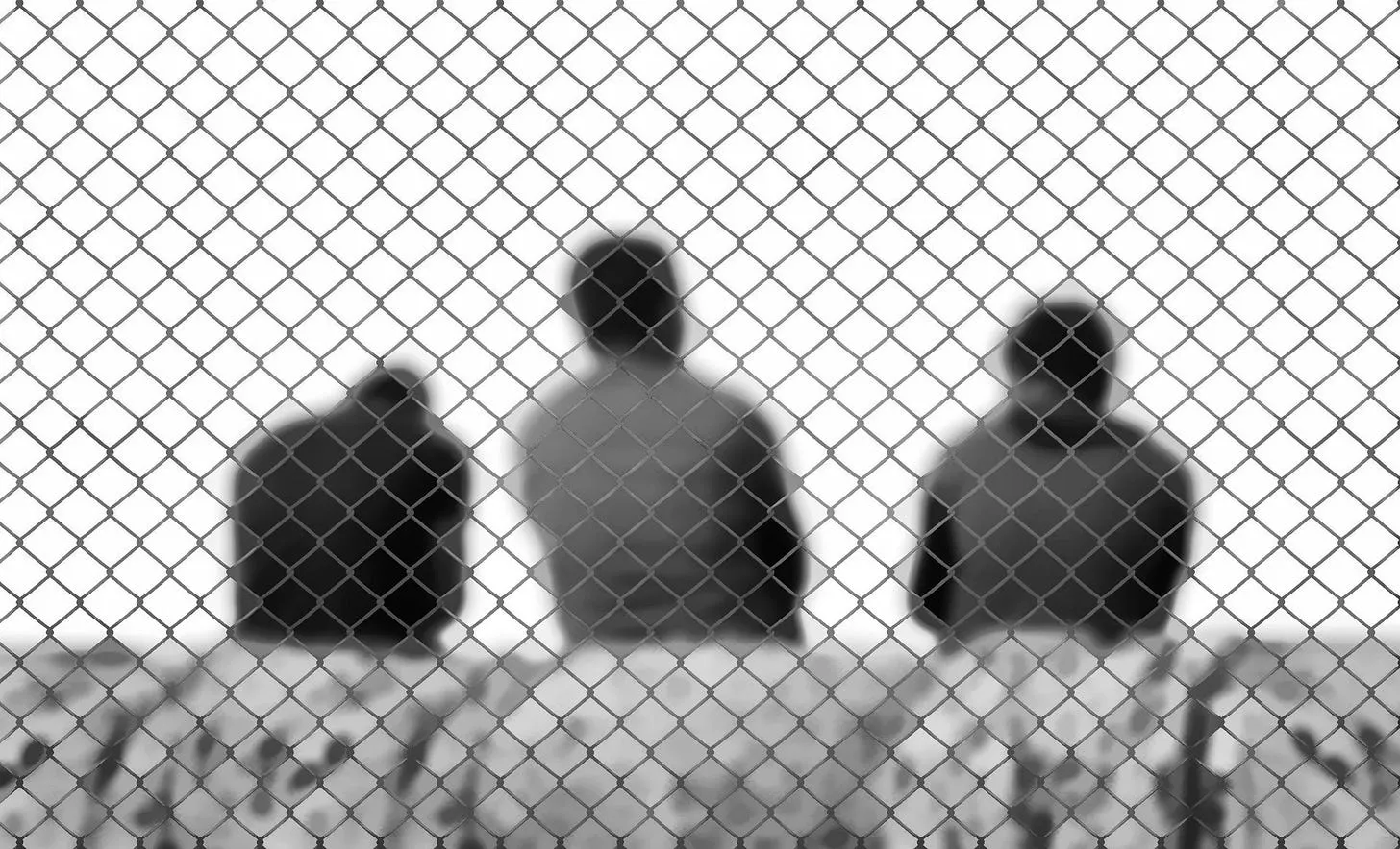 Migranten sitzen mit dem Rücken zur Kamera hinter einem Maschendrahtzaun. Stilisiert in schwarz-weiß. Foto: Pixabay.com.