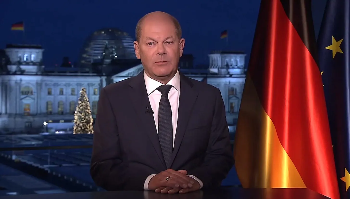 Bundeskanzler Olaf Scholz bei seiner Neujahrsansprache 2022. Er sitzt mit übereinandergefalteten Händen an einem Tisch. Im Hintergrund der Bundestag bei Nacht mit einem beleuchteten Weihnachtsbaum. Rechts neben ihm die Deutschlandflagge und eine Flagge der Europäischen Union.