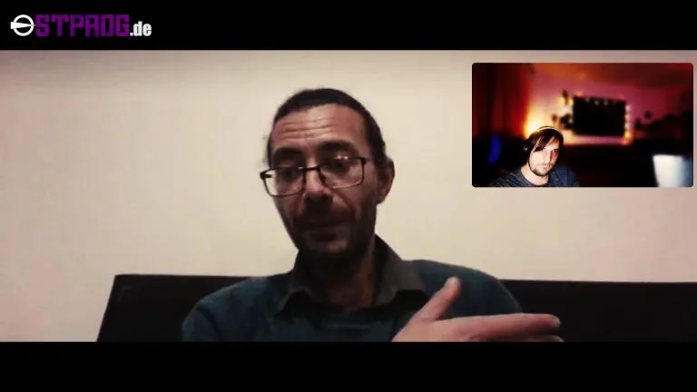 Karam Bales auf einem Sofa vor einer weißen Wand. Rechts oben im Bild ist Gunnar Hamann zu sehen in einem kleinen Fenster in einem bunt beleuchteten Zimmer. Oben links auf dem Screenshot steht Ostprog.de.