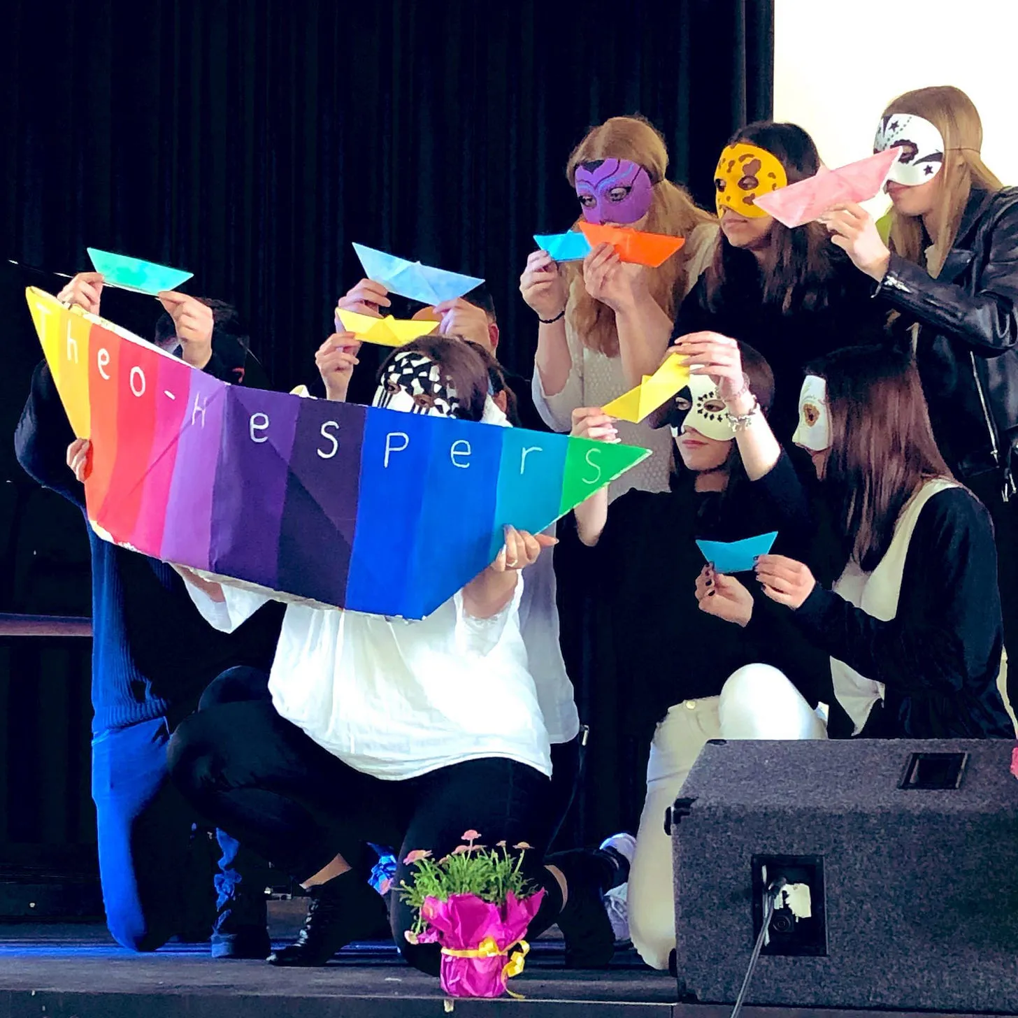 Schülerinnen und Schüler mit fantasievollen Masken halten ein großes Papierschiff in Regenbogenfarben auf dem Theo Hespers steht