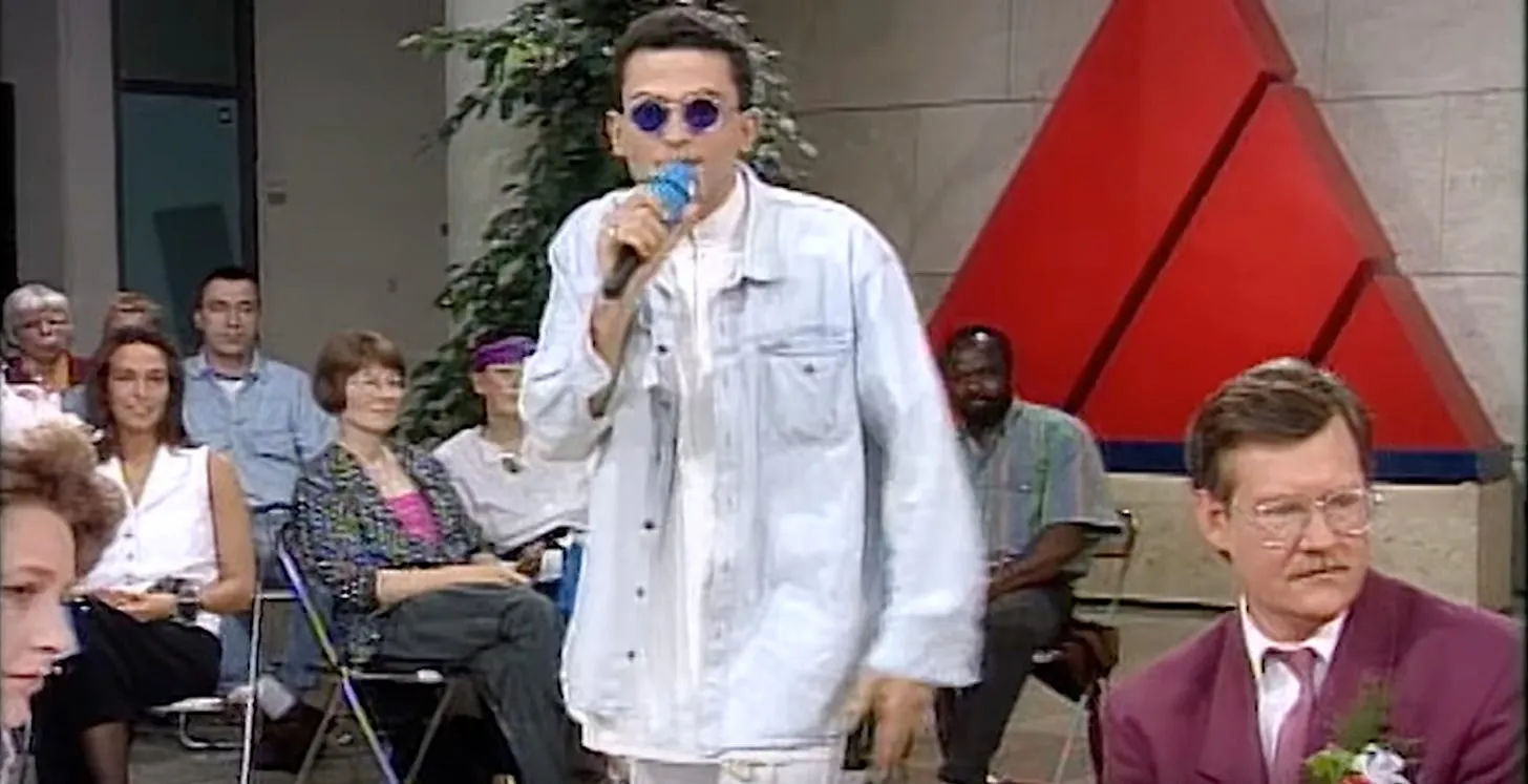 Fatih Çevikkollu mit runder Sonnenbrille mit blauen Gläsern und goldenem Rahmen, hellem Jeanshemd, Mikrofon, im Hintergrund das rote Dreieck der Sendung Aktuelle Stunde des WDR