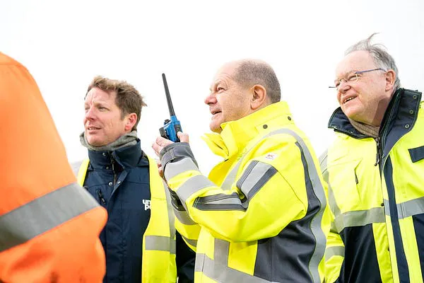 Bundeskanzler Olaf Scholz besichtigt das LNG-Terminal in Wilhelmshaven. Er hat eine gelbe Warnjacke an und hält ein Funkgerät in der Hand. Daneben ist Niedersachsens Ministerpräsident, Stephan Weil, zu sehen.